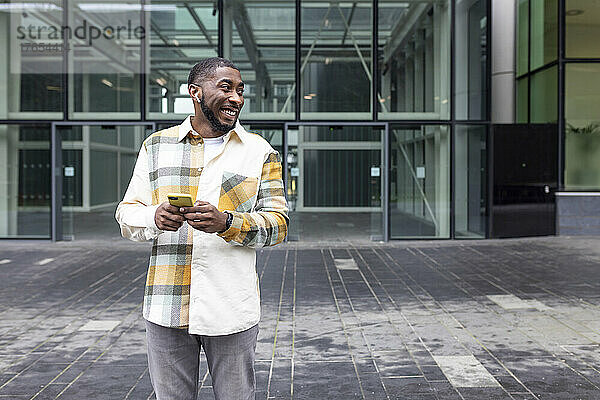 Lächelnder Mann mit Mobiltelefon steht vor einem Glasgebäude