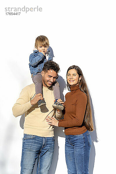 Glücklicher Mann trägt Sohn auf der Schulter und steht neben einer jungen Frau vor einer weißen Wand