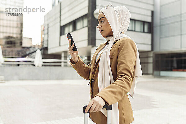Geschäftsfrau mit Hijab surft über ihr Smartphone im Netz und steht vor einem modernen Gebäude