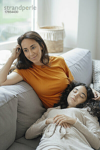 Teenage girl resting in lap of thoughtful mother on sofa at home