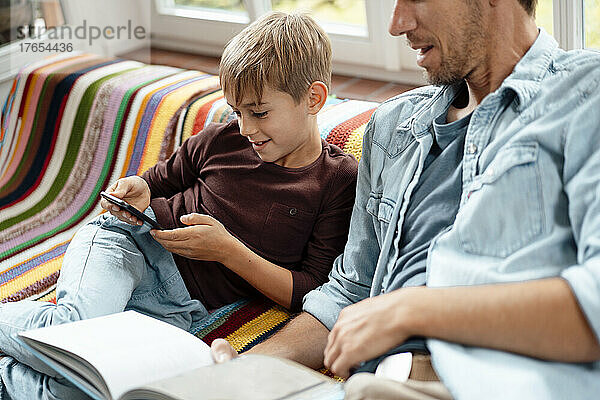 Mann mit Buch blickt Sohn mit Smartphone an  der zu Hause auf dem Sofa sitzt