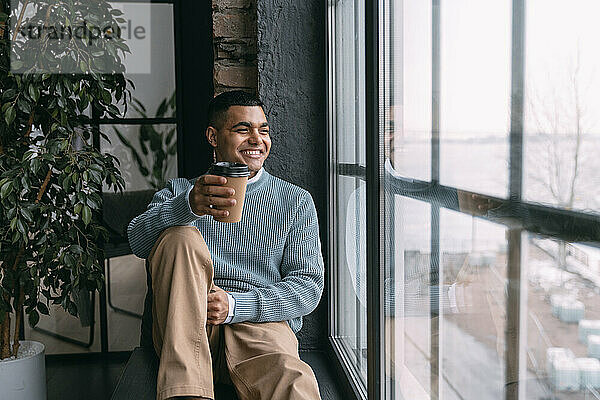 Glücklicher junger Mann mit Einwegkaffeetasse  der zu Hause durch das Fenster schaut
