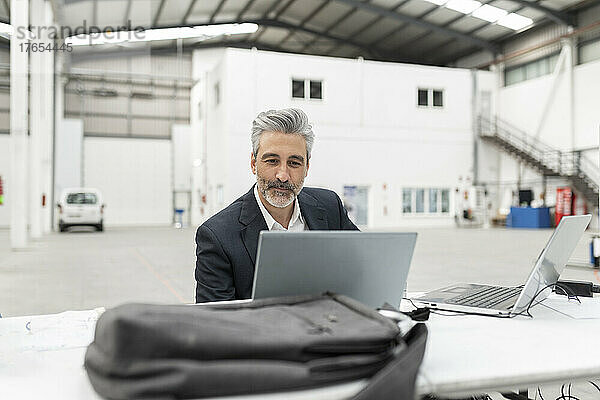 Geschäftsmann mit Laptop sitzt am Schreibtisch in der Fabrik