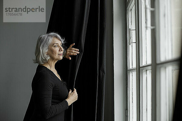Ältere Frau steht am Vorhang und blickt zu Hause durch das Fenster