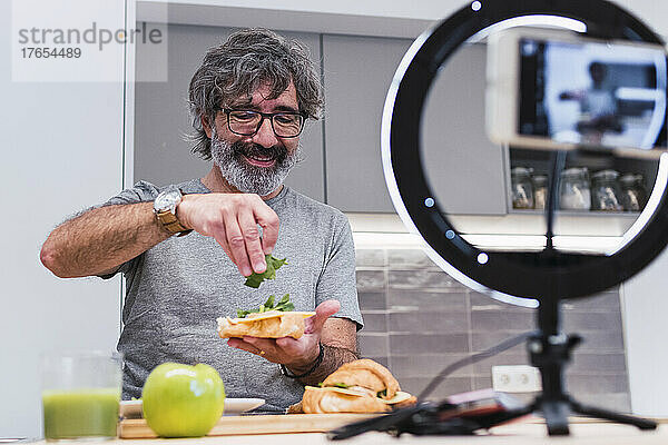 Glücklicher älterer Vlogger  der zu Hause auf der Kücheninsel Sandwich mit Croissant zubereitet