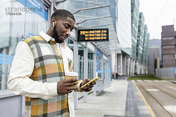 Mann hält Einwegbecher in der Hand und benutzt Mobiltelefon an Straßenbahnhaltestelle