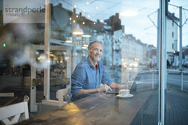 Lächelnder Freiberufler mit Laptop durch Glasfenster im Café gesehen