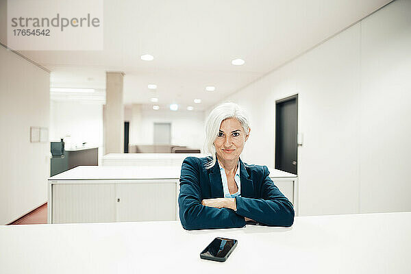 Geschäftsfrau sitzt mit verschränkten Armen am Schreibtisch im Büro