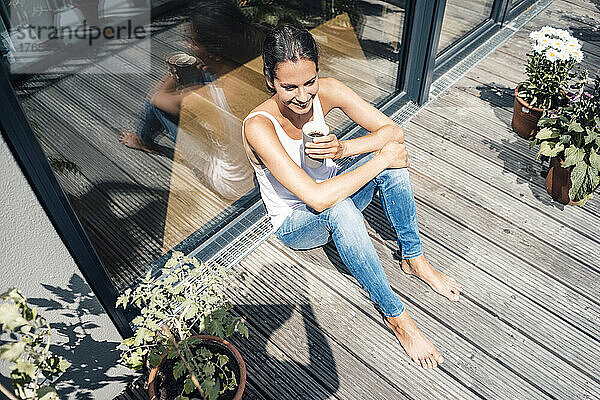 Glückliche Frau mit Kaffeetasse sitzt an einem sonnigen Tag auf dem Balkon