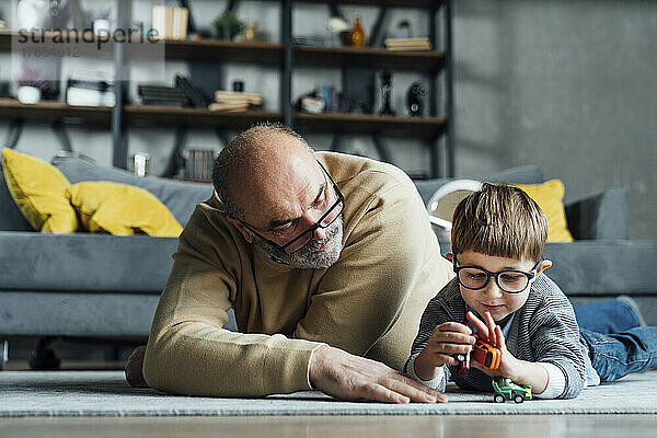 Älterer Mann schaut Enkel an  der im Wohnzimmer mit Spielzeugauto spielt
