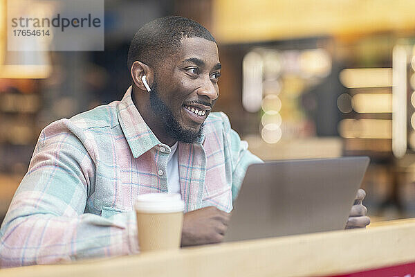 Lächelnder Mann mit Laptop sitzt im Café und sieht durch Glas