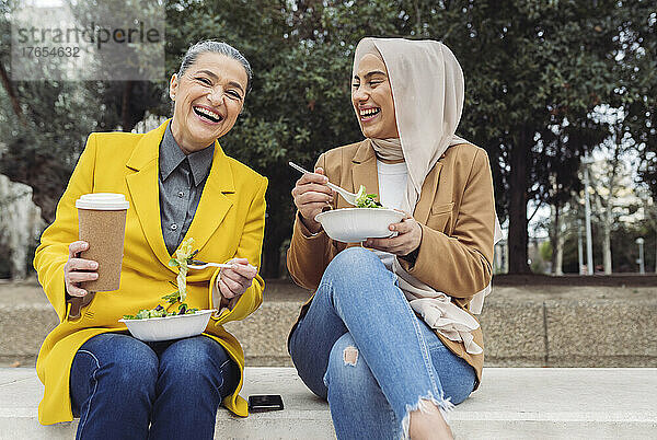 Fröhliche Geschäftsfrauen mit Salatschüsseln und Einwegkaffeetasse sitzen auf Stufen