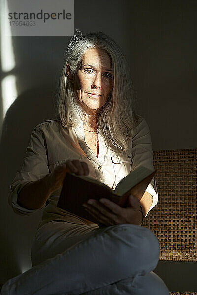 Frau mit grauen Haaren hält ein Buch in der Hand und sitzt zu Hause auf einem Stuhl