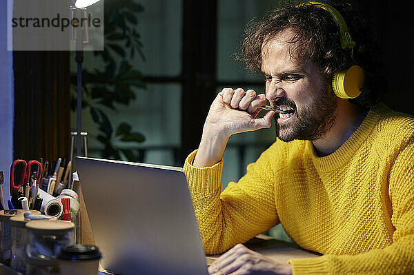 Geschäftsmann mit Laptop beißt Stift und hört Musik über Kopfhörer im Büro
