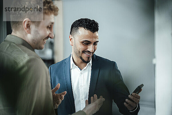 Lächelnder Geschäftsmann zeigt seinem Kollegen im Büro sein Mobiltelefon
