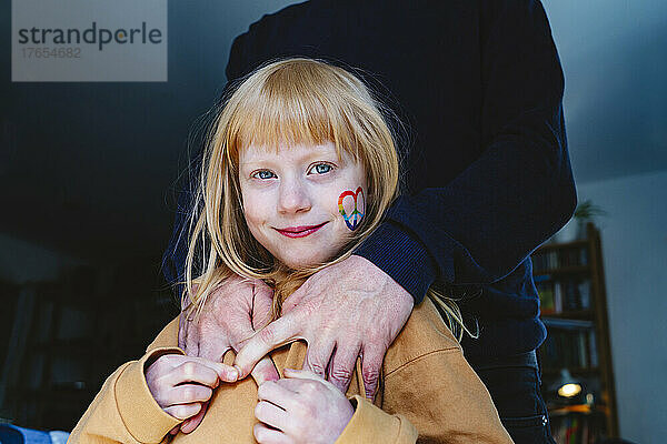 Lächelndes blondes Mädchen mit Friedensaufkleber auf der Wange  das mit seinem Vater zu Hause steht
