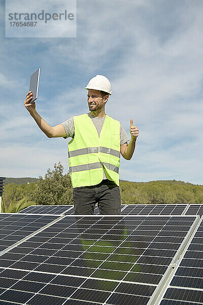 Ingenieur macht Selfie mit erhobenem Daumen inmitten von Sonnenkollektoren auf dem Dach