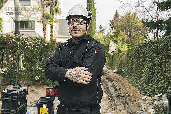 Lächelnder Bauarbeiter steht mit verschränkten Armen auf der Baustelle