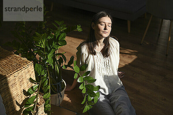 Frau mit geschlossenen Augen genießt das Sonnenlicht  während sie zu Hause neben einer Zimmerpflanze sitzt
