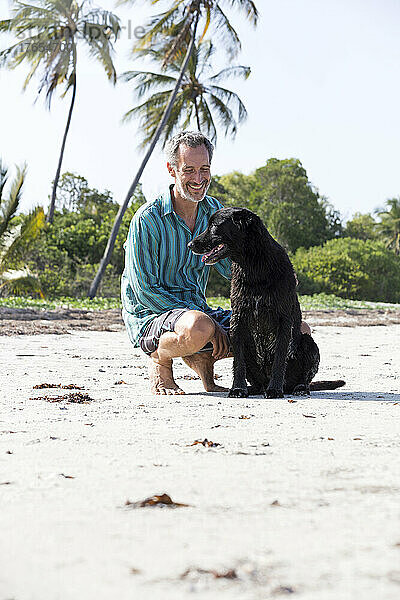Lächelnder Mann kauert an einem sonnigen Tag neben einem Hund am Strand
