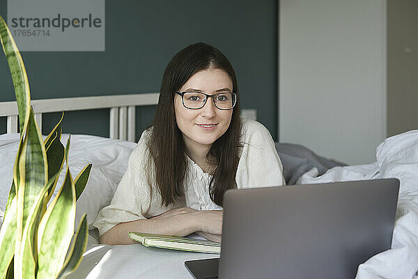 Lächelnde Frau mit Brille und Laptop liegt zu Hause im Bett