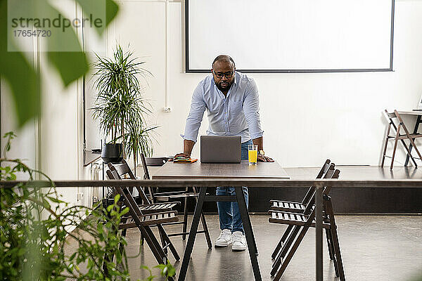 Selbstbewusster Geschäftsmann mit Laptop  der im Büro auf dem Schreibtisch lehnt