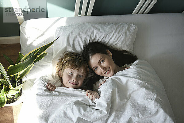 Lächelnde Mutter und Sohn mit Decke liegen zu Hause im Bett