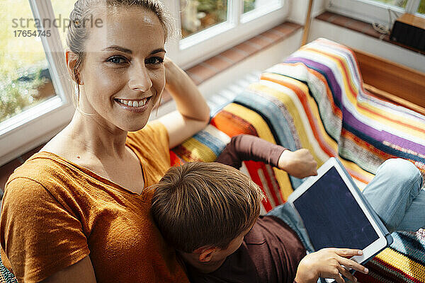 Lächelnde Mutter sitzt neben ihrem Sohn und benutzt Tablet-PC auf dem heimischen Sofa