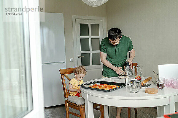 Mann bereitet Pizza zu  während seine Tochter auf einem Stuhl in der Küche sitzt
