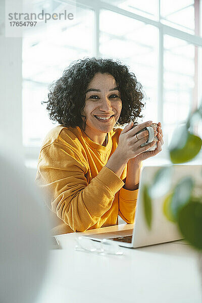 Glückliche Frau mit Kaffeetasse und Laptop am Tisch