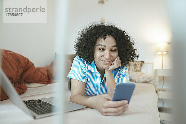 Lächelnde Frau mit Laptop und Smartphone im heimischen Bett