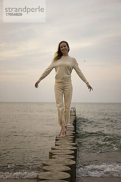 Glückliche Frau mit erhobenen Armen  die auf einem Holzpfosten am Strand spaziert