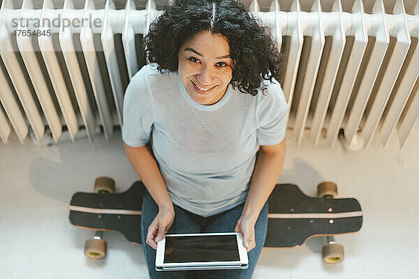 Glückliche Frau sitzt zu Hause auf Skateboard mit Tablet-PC