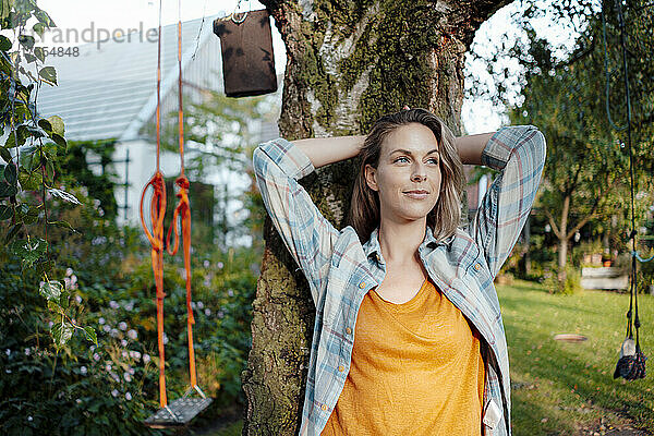 Lächelnde Frau steht mit den Händen hinter dem Kopf vor einem Baumstamm im Hinterhof