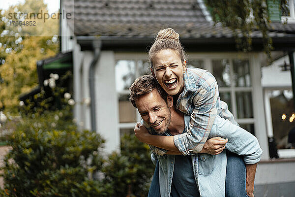 Lächelnder Mann gibt seiner fröhlichen Freundin eine Huckepackfahrt  die es im Hinterhof genießt