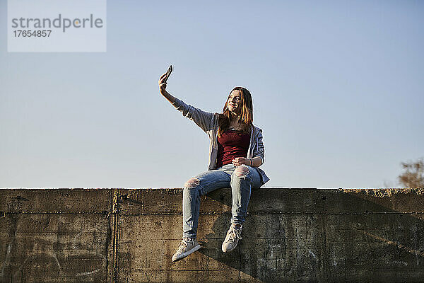 Junge Frau sitzt auf Betonwand und macht ein Selfie