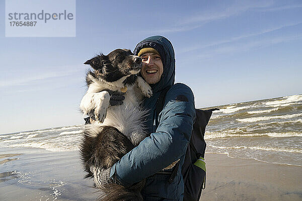 Glücklicher junger Mann  der an einem sonnigen Tag seinen Hund am Strand trägt