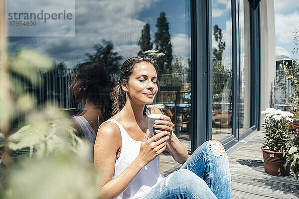 Lächelnde Frau mit geschlossenen Augen und einer Einweg-Kaffeetasse  die vor einem Glasfenster auf dem Balkon sitzt