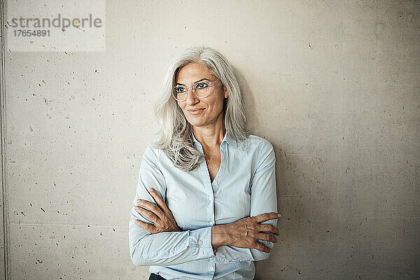 Lächelnde Geschäftsfrau mit Brille steht mit verschränkten Armen vor der Wand