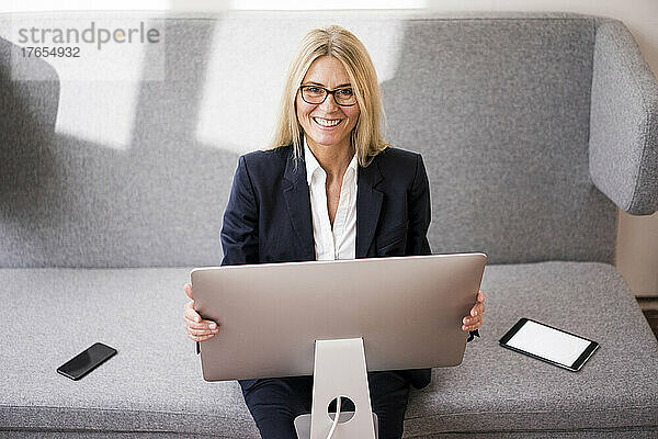 Lächelnde Geschäftsfrau mit Computer sitzt auf dem Sofa im Büro