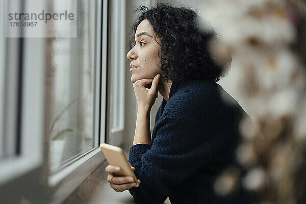 Nachdenkliche Frau mit Smartphone  die durch das Fenster schaut