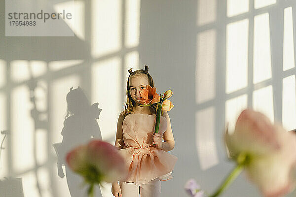 Mädchen hält Blume im Gesicht und steht vor einer weißen Wand