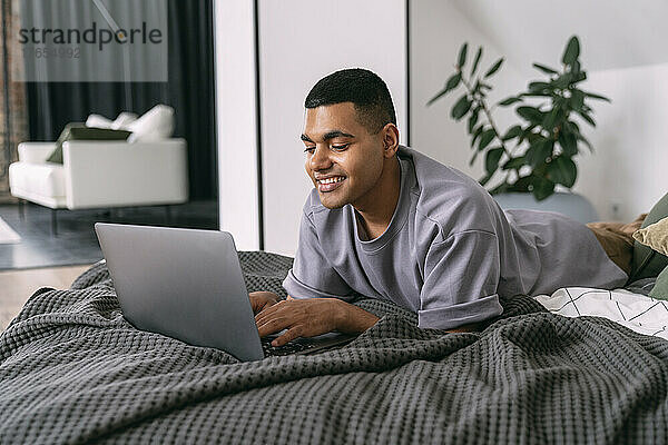 Lächelnder junger Mann mit Laptop  der zu Hause im Bett liegt