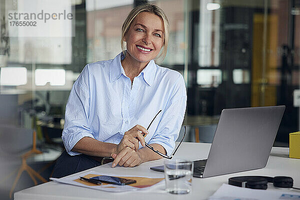 Lächelnde Geschäftsfrau mit Brille und Laptop am Schreibtisch im Büro