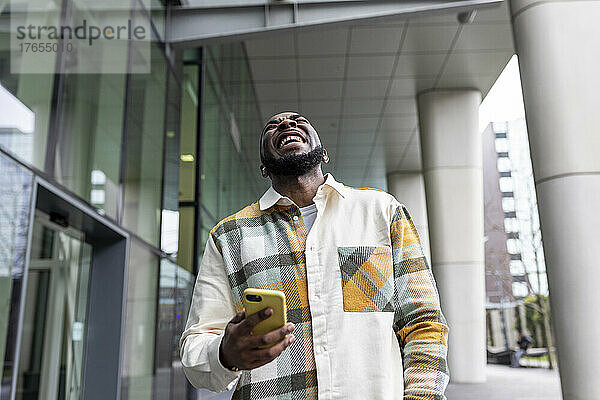 Fröhlicher Mann hält Smartphone und lacht vor Glasgebäude