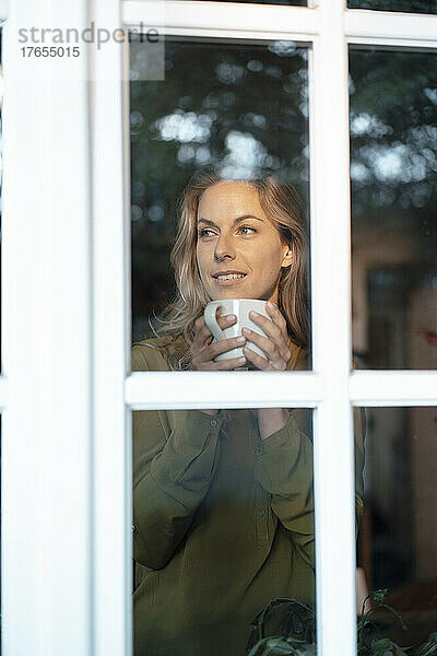 Beautiful blond woman holding coffee mug seen through glass window