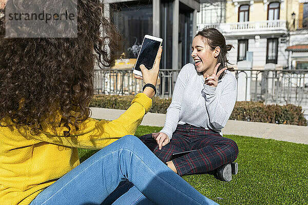 Junge Frau fotografiert im Park einen Freund  der per Smartphone ein Friedenszeichen zeigt