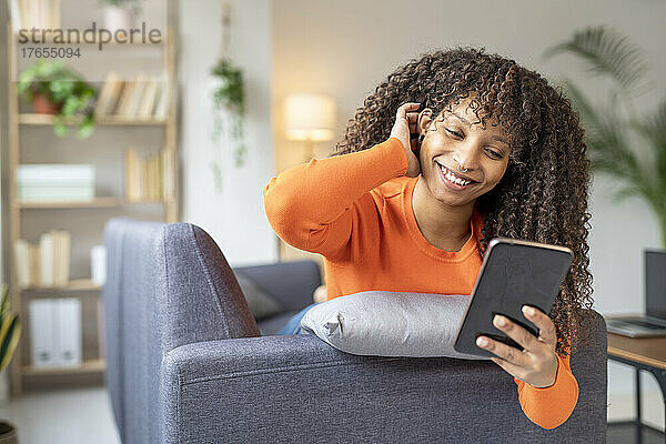Glückliche junge Frau mit der Hand im Haar  die zu Hause auf dem Sofa ihr Mobiltelefon benutzt
