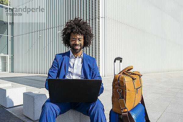Glücklicher Geschäftsmann  der an einem sonnigen Tag am Gepäck sitzt und seinen Laptop benutzt