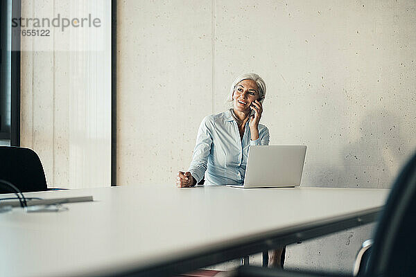 Lächelnde Geschäftsfrau  die mit einem Laptop am Schreibtisch sitzt und mit einem Smartphone spricht